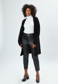 Cardigan nero a maglia sopra una dolcevita bianca, abbinato a pantaloni neri in ecopelle e scarpe con tacco nere. Texture morbide, design aderente.