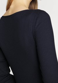 Nahaufnahme des oberen Rückens und Nackens einer Person, die ein dunkelblaues, geripptes Langarmoberteil vor einem neutralen Hintergrund trägt.