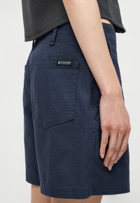 Άτομο που φορά μπλε σκούρο σορτς με πίσω τσέπη και ετικέτα της Columbia Sportswear Company, στέκεται με το δεξί του χέρι χαλαρό δίπλα στο σώμα.