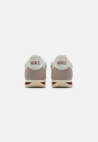 Nike sportcipők világosbarna velúrból készült felsőrésszel, fehér saroktabbal, piros logóval, valamint texturált krémszínű talppal, bordó díszcsíkkal.