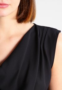 Robe noire avec un décolleté drapé et des manches courtes. Le tissu est lisse, avec un plissage subtil près de l'épaule, créant de doux plis.