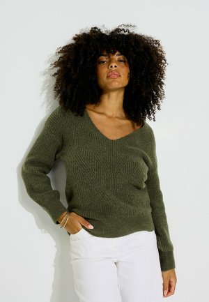 Mujer con cabello rizado que lleva un suéter verde texturizado con cuello en V y pantalones blancos, de pie con una mano en el bolsillo contra un fondo liso.