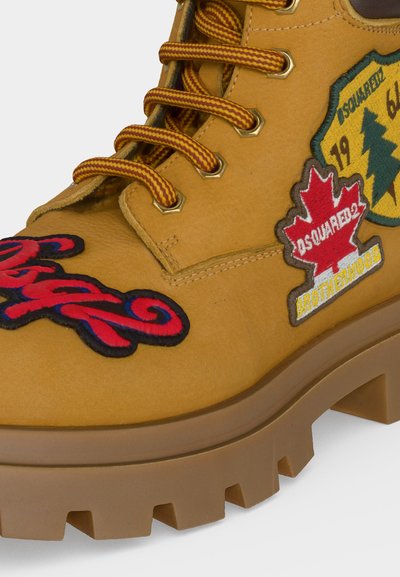 Botte jaune à lacets avec semelle épaisse, décorée de patchs brodés rouges et verts représentant une feuille d'érable, du texte et un emblème de pin.