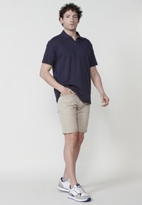 Polo shirt marine, à manches courtes, associé à un short beige clair, avec des bords roulés. Chaussures : baskets blanches avec des accents bleu marine et rouge.