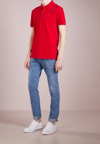 Polo rouge avec un petit logo, associé à un jean bleu clair et des baskets blanches. Tissu lisse avec un col classique et des manches courtes.