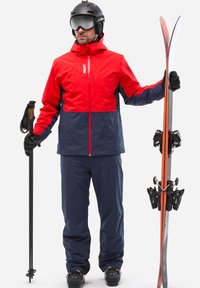Rote und marineblaue Skijacke, marineblaue Hose, schwarze Handschuhe und Helm. Hält Skier mit schwarzen Bindungen und einen Skistock, vor einem weißen Hintergrund.