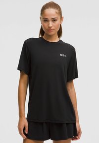 Fiatal nő, copfba kötve, laza fekete pólót és azzal megegyező rövidnadrágot viselve, egy sima világos háttér előtt áll.