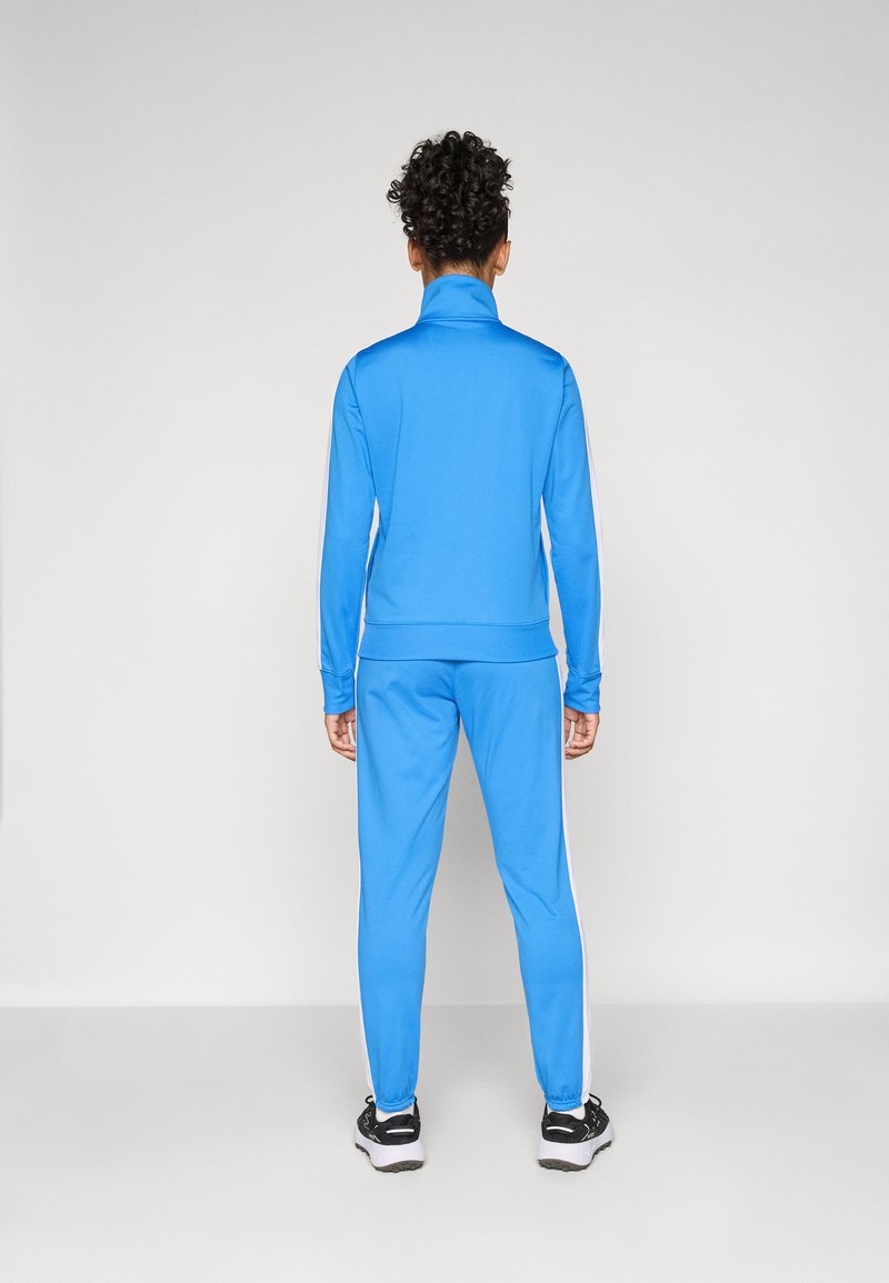 Chándal deportivo azul que incluye una chaqueta con cremallera y pantalones con rayas blancas a los lados. Tejido suave con silueta ajustada. Zapatillas negras incluidas.