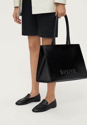 Person, der holder en stor sort læder tote taske med "kazar" logo, iført sorte shorts, beige jakke og sorte loafers.