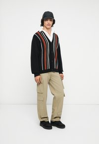 Zwarte gebreide cardigan met gekleurde verticale strepen, knoopsluiting, gecombineerd met beige cargobroek en zwarte sneakers, gedragen met een pet.