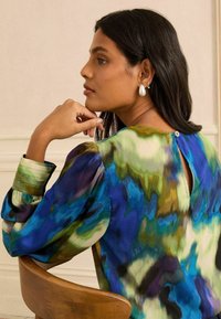 Többszínű blúz kék, zöld és lila színekben, fodros ujjakkal, kulcslyuk hátlappal gombbal, és selymes textúrával.
