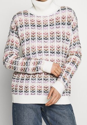Pull à col roulé blanc avec motif chevron multicolore tricoté, porté avec un jean bleu, mains ajustant la manche.