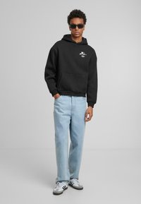 Svart hettegenser med frontlomme og brodert design, parret med lyseblå straight-leg jeans og svart-hvite sneakers.
