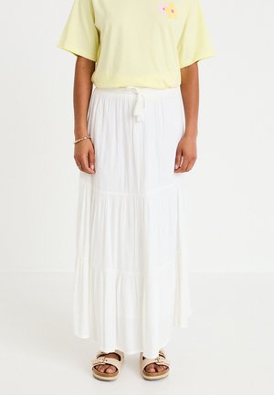 Woman wearing a yellow short-sleeve shirt, long white tiered skirt with drawstring, and beige sandals standing against a white background.