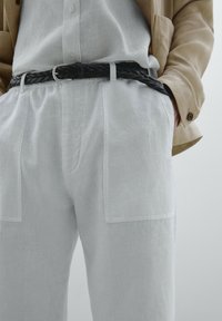 Pantalons en lin blancs avec poches avant, en tissu texturé, combinés avec une ceinture tissée noire et une chemise boutonnée gris clair.