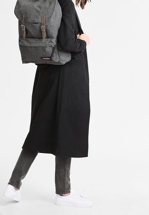 Henkilö, jolla on pitkä musta takki, harmaat farkut, valkoiset lenkkarit ja kantaa leveillä hihnoilla varustettua isoa harmaata reppua toisella olkapäällä.