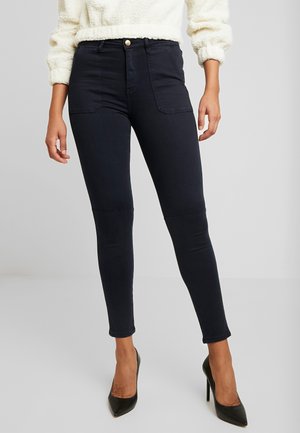 Frau trägt hoch taillierte schwarze Skinny-Jeans mit vorderen aufgesetzten Taschen, kombiniert mit schwarzen spitz zulaufenden High Heels und einem weißen strukturierten Pullover.