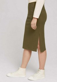 Jupe crayon mi-longue couleur vert olive en tissu texturé, avec une fente latérale, associée à des baskets montantes blanches en toile.