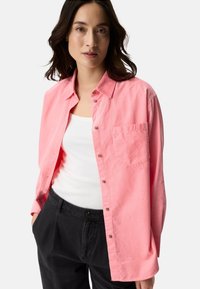 Rosa Hemd mit Knöpfen, Kragen, einer Brusttasche und langen Ärmeln, kombiniert mit einem weißen Tanktop und schwarzen Hosen.