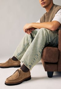 Brune ruskindsko med rundet tå og snøredetalje, båret med lyseblå cowboybukser og en strikket beige vest. Tekstureret brun lænestol.