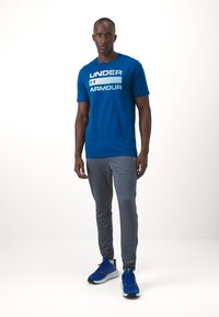Muž stojaci oblečený v modrom tričku Under Armour, šedých športových nohaviciach a modrých bežeckých topánkach na jednoduchom bielom pozadí.