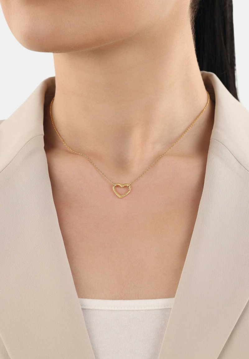Collana a pendente a forma di cuore dorato attorno al collo di una donna, che indossa un blazer beige e una maglietta bianca, su uno sfondo chiaro e semplice.