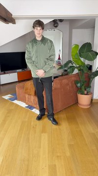 Lett olivengrønn jakke med innstrammet midje, svarte bukser og svarte sko, står i et rom med tregulv og planter.