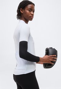 Άτομο με πλεξούδες στα μαλλιά, φορώντας λευκή φανέλα ποδηλασίας και μαύρα μανίκια στα χέρια, κρατώντας ένα μαύρο κράνος ποδηλάτου.