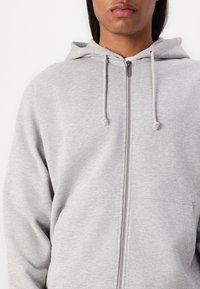 Svetlošedý zipsový hoodie vyrobený z mäkkej tkaniny. Má prednú kapsu a kapucňu so šnúrkami. Hladká textura s uvoľneným strihom.