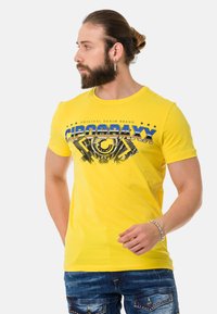 Yellow cotton t-shirt featuring a graphic print in blue and black, rounded neckline, and short sleeves. Worn with denim pants.