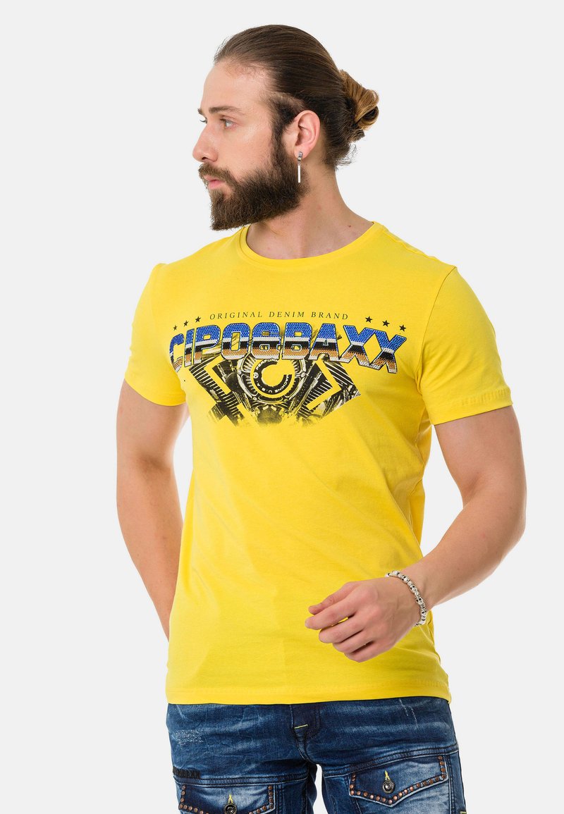Yellow cotton t-shirt featuring a graphic print in blue and black, rounded neckline, and short sleeves. Worn with denim pants.