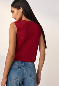 Top rosso senza maniche con design aderente, tessuto strutturato e lunghezza leggermente corta, abbinato a jeans di denim blu a vita alta.