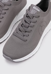 Zapatillas deportivas grises con una parte superior de malla texturizada, suela blanca y cordones planos. Presentan un logo discreto en el lateral y un collar acolchado.