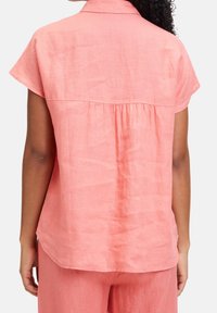 Nainen, joka pukeutuu löysään, lyhythihaiseen korallinpunertavaan puseroon ja siihen sopiviin housuihin, kuvattuna takaa valkoista taustaa vasten.