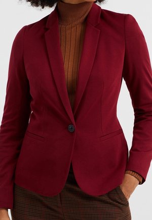 Kobieta ubrana w dopasowaną, ciemnoczerwoną marynarkę z jednym czarnym guzikiem, na brązowym prążkowanym golfie i brązowych spodniach w kratkę, z ręką w kieszeni.