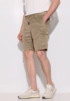 Mand iført beige kordfløjls cargo-shorts og hvide sneakers, stående mod en ensfarvet lys baggrund.