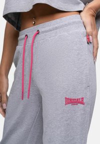 Grå svettbyxor med rosa snörning, med en röd Lonsdale-logotyp. Tillverkade av mjukt tyg, de har sidofickor och en accent med den brittiska flaggan.