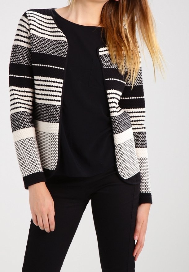 Femme portant un cardigan en maille à motifs noir et blanc sur un haut noir avec un pantalon noir, debout devant un fond uni.