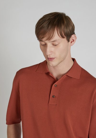 Polo à manches courtes de couleur rouille en tissu texturé, avec un col et deux boutons foncés sur la patte de boutonnage.
