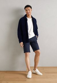Marineblå kordfløyelsjakke, hvit t-skjorte, marineblå shorts, og hvite joggesko. Avslappet passform med teksturert stoff og enkelt design.