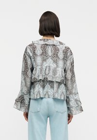 Femme vue de dos portant un chemisier à manches longues transparent à motif, avec des volants superposés, et un jean taille haute bleu clair.