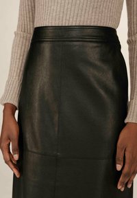 Gros plan sur une personne portant une jupe en cuir noire et un pull à manches longues beige à côtes, montrant des mains détendues sur les côtés.