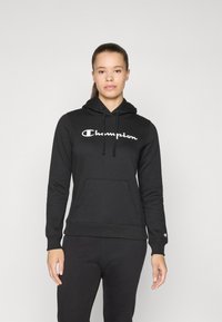 Svart bomullshoodie med frontlomme og snor. Den har en hvit "Champion"-logo på brystet og matchende svarte bukser.