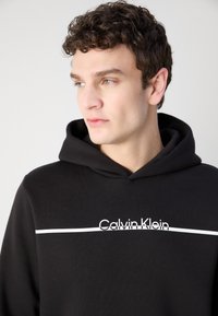 Črna kapuca iz bombaža z belo črto spredaj in logotipom "Calvin Klein". Ima kapuco s vrvico in rebraste manšete.