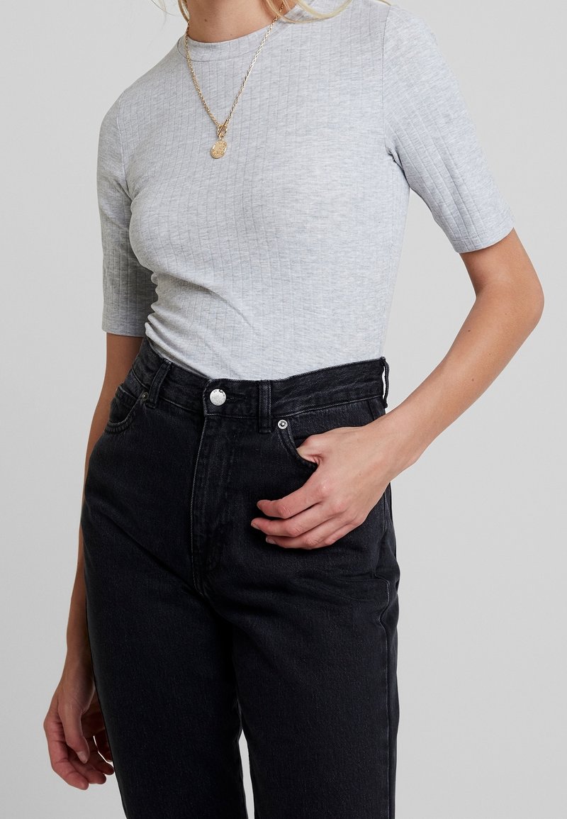 Femme portant un haut à manches courtes côtelé gris clair rentré dans un jean noir taille haute, avec la main dans la poche et un collier en or.