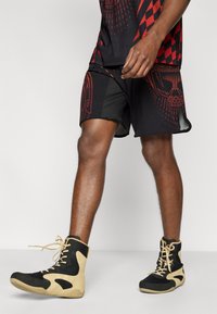 Μαύρα αθλητικά σορτς με κόκκινα γραφικά μοτίβα και μαύρες λεπτομέρειες, σε συνδυασμό με ψηλά παπούτσια που έχουν δίχτυ επάνω μέρος και καφέ λεπτομέρειες.