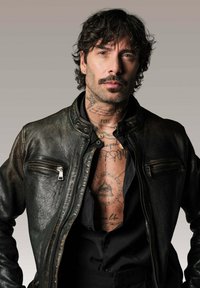 Veste de moto en cuir noir avec quincaillerie en métal argenté et deux poches zippées à l'avant. La personne arbore des tatouages et porte un t-shirt noir en dessous.