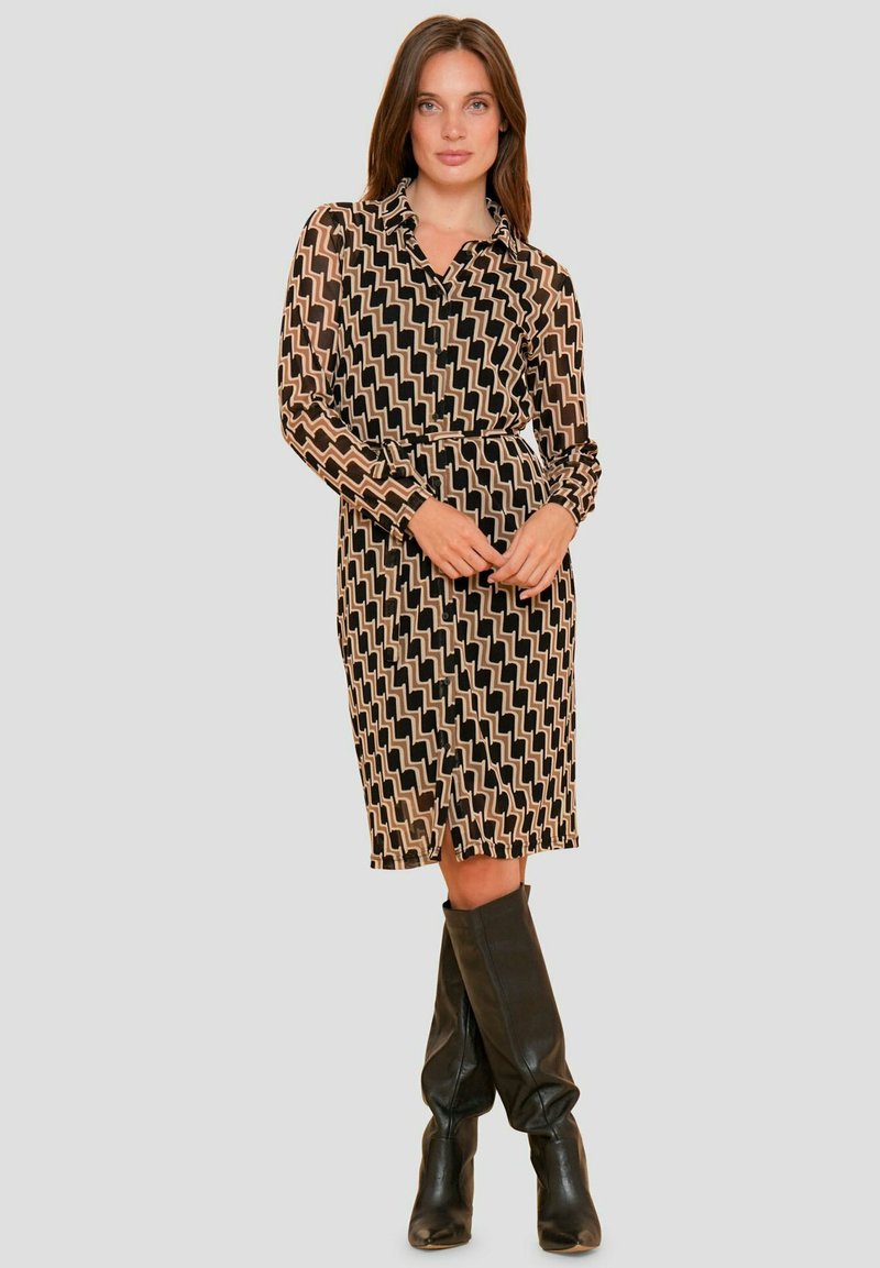 Langarmige jurk in zwart en beige zigzagpatroon, met een kraag, een ceintuur in de taille en een knielange snit, gecombineerd met zwarte laarzen tot de knie.