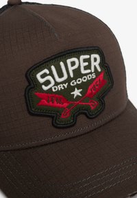 Gorra de béisbol marrón con un parche bordado en relieve que lleva el texto "SUPER DRY GOODS" en blanco, rodeado de acentos rojos y detalles en verde.