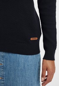 Navy gestrickter Pullover mit strukturiertem Muster und gerippten Bündchen; enthält ein kleines braunes Lederetikett mit dem Markennamen "Oxmo" am Saum.